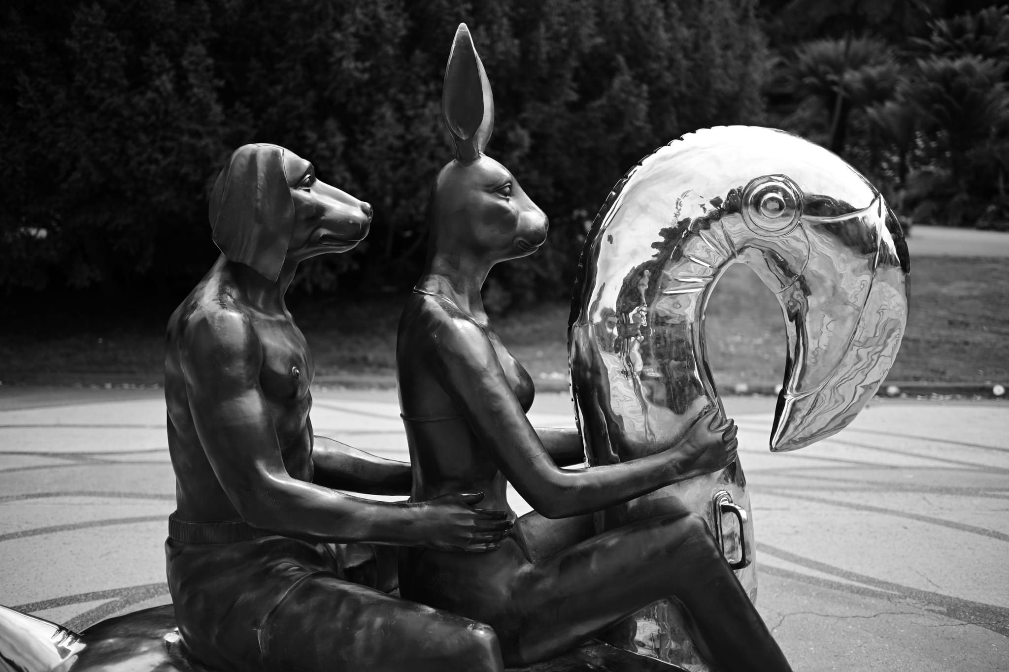 A black and white sculpture of two anthropomorphic figures, one with a dog head and the other with a rabbit head, sitting on a motorcycle with a large, reflective balloon-like object in front