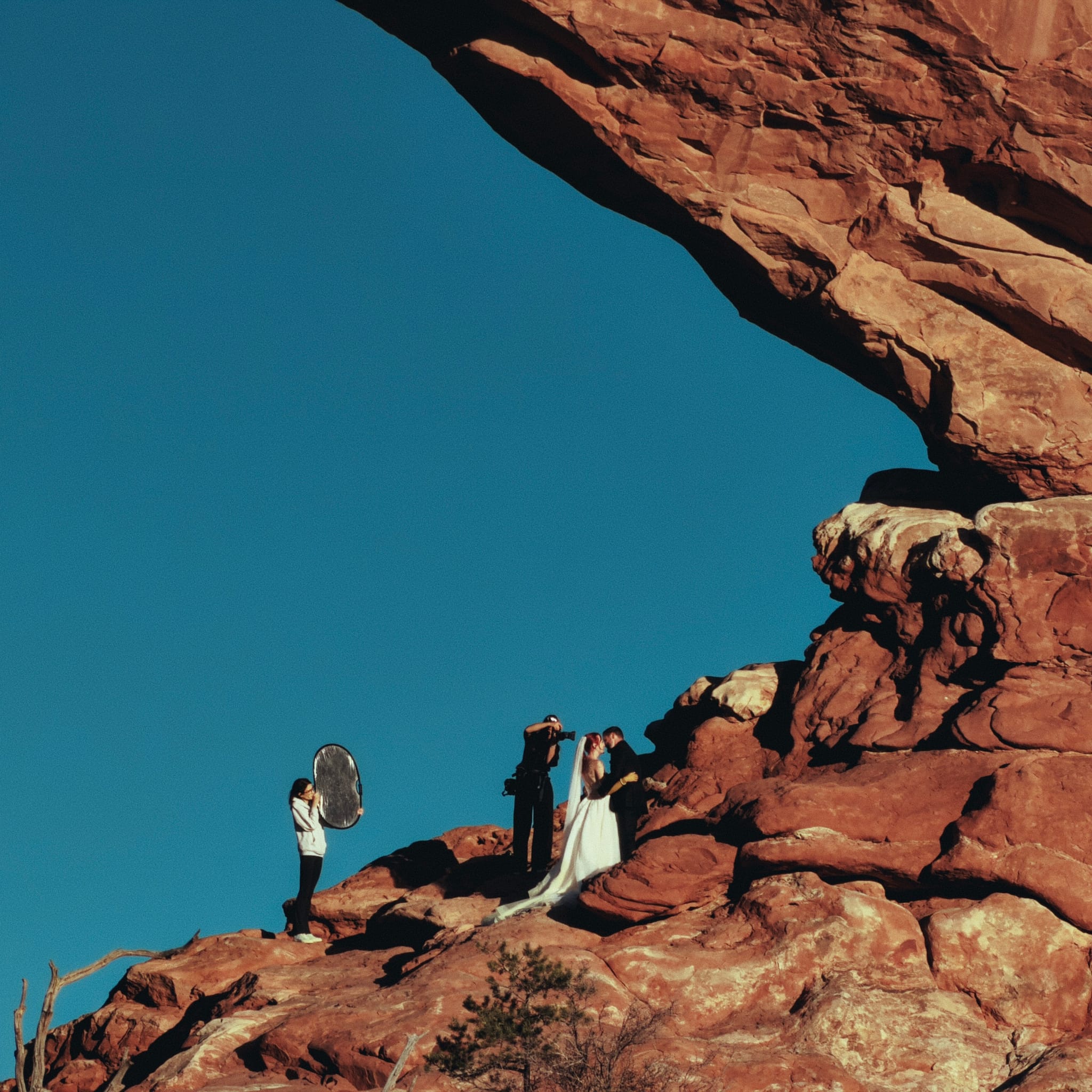 Wedding couple and photo crew on red sandstone ledge beneath a large natural arch against clear blue sky