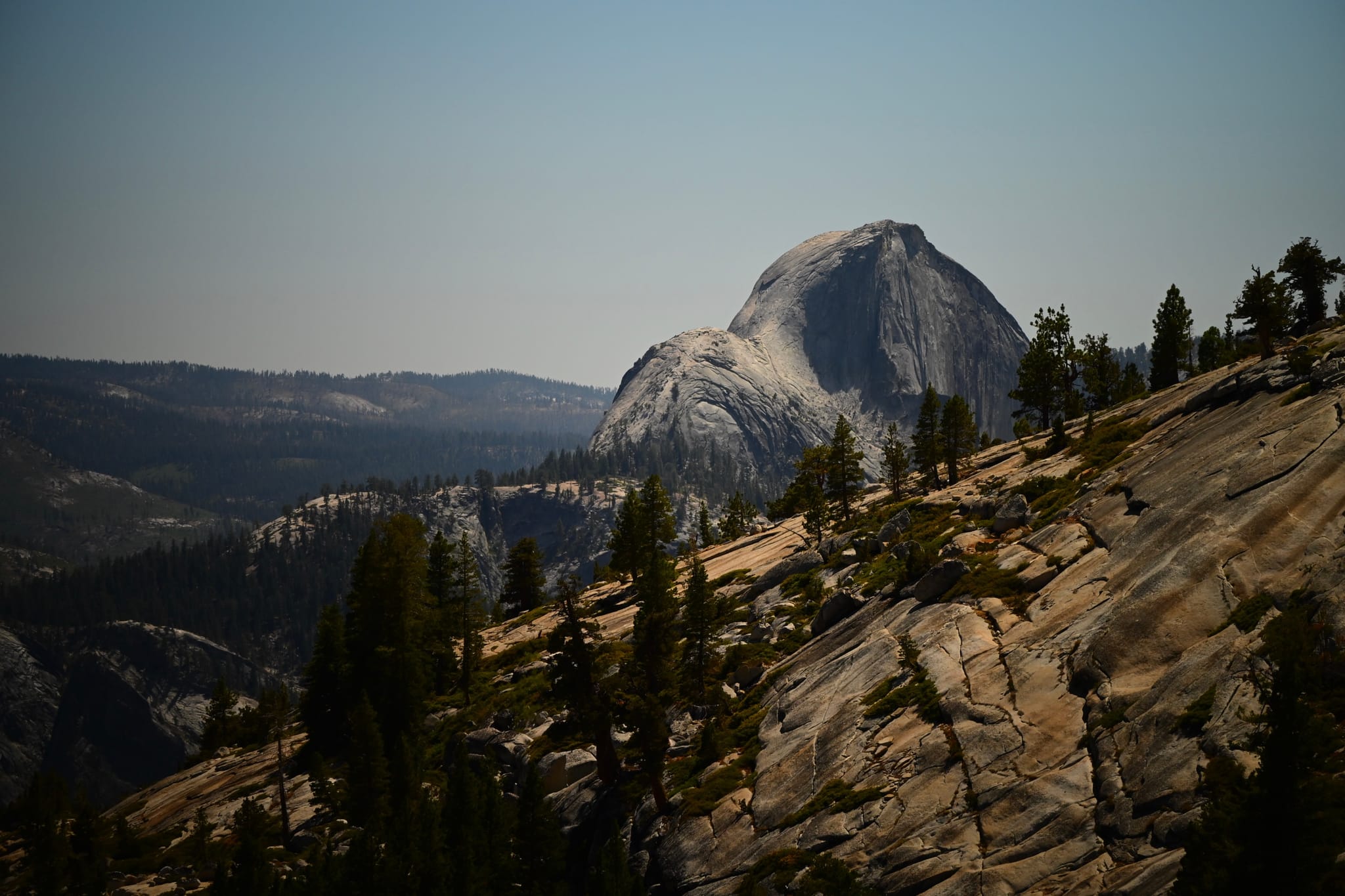 A mountainous landscape with a prominent granite dome in the background, surrounded by trees and rocky terrain under a clear sky