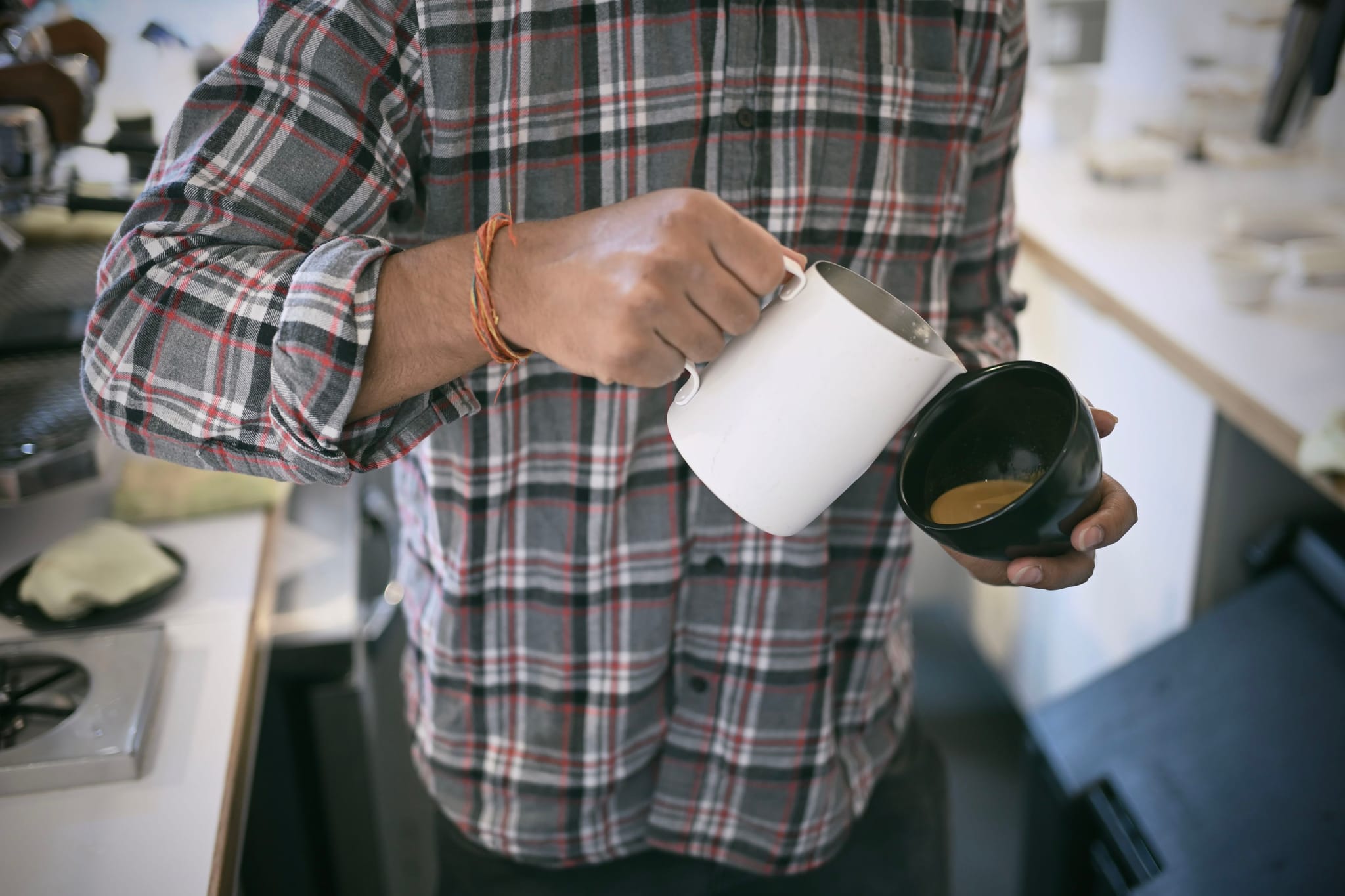 A person in a plaid shirt pouring milk from a white pitcher into a cup of coffee