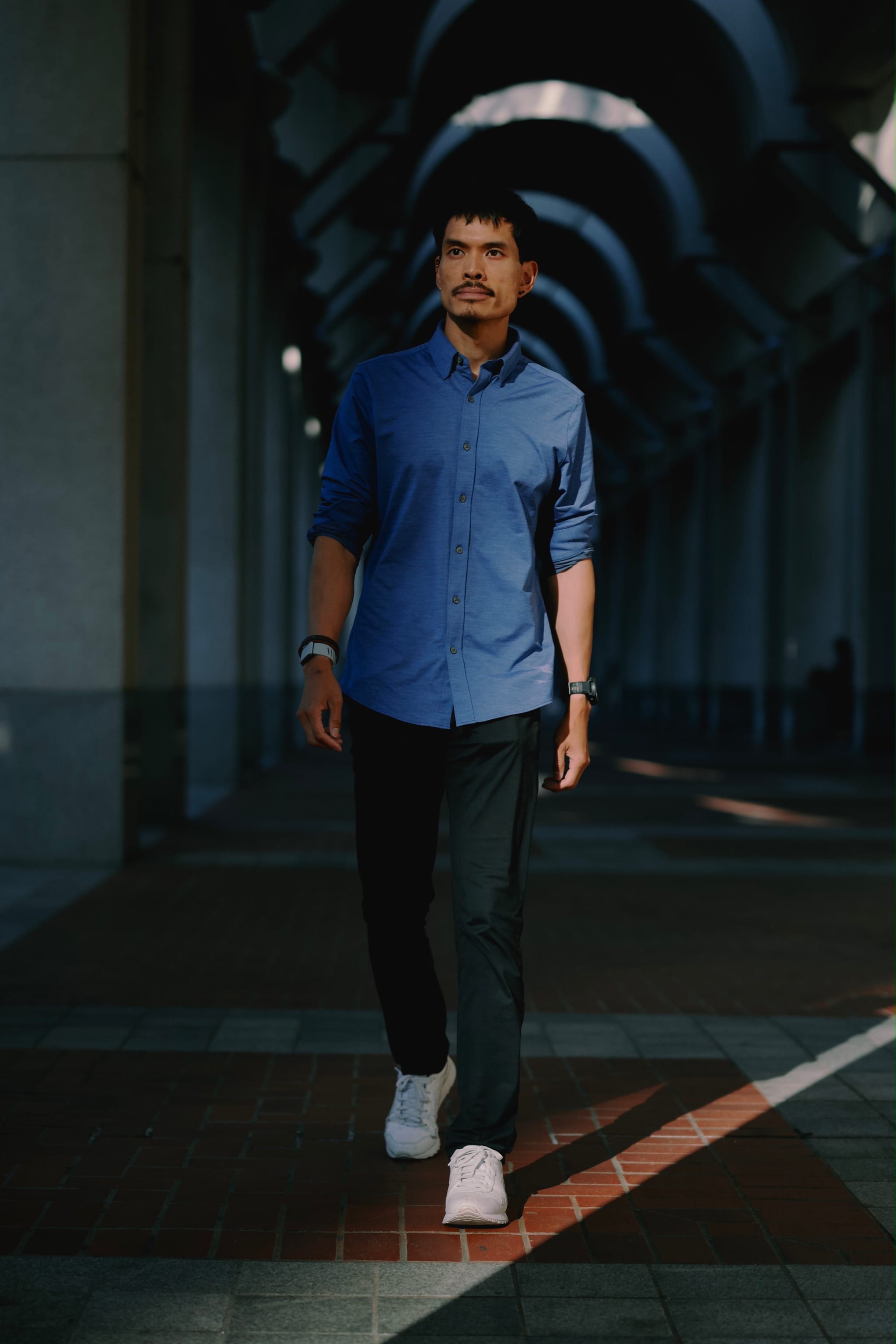 A man walks confidently through a shaded archway corridor, dressed in a blue shirt and dark pants, illuminated by natural light