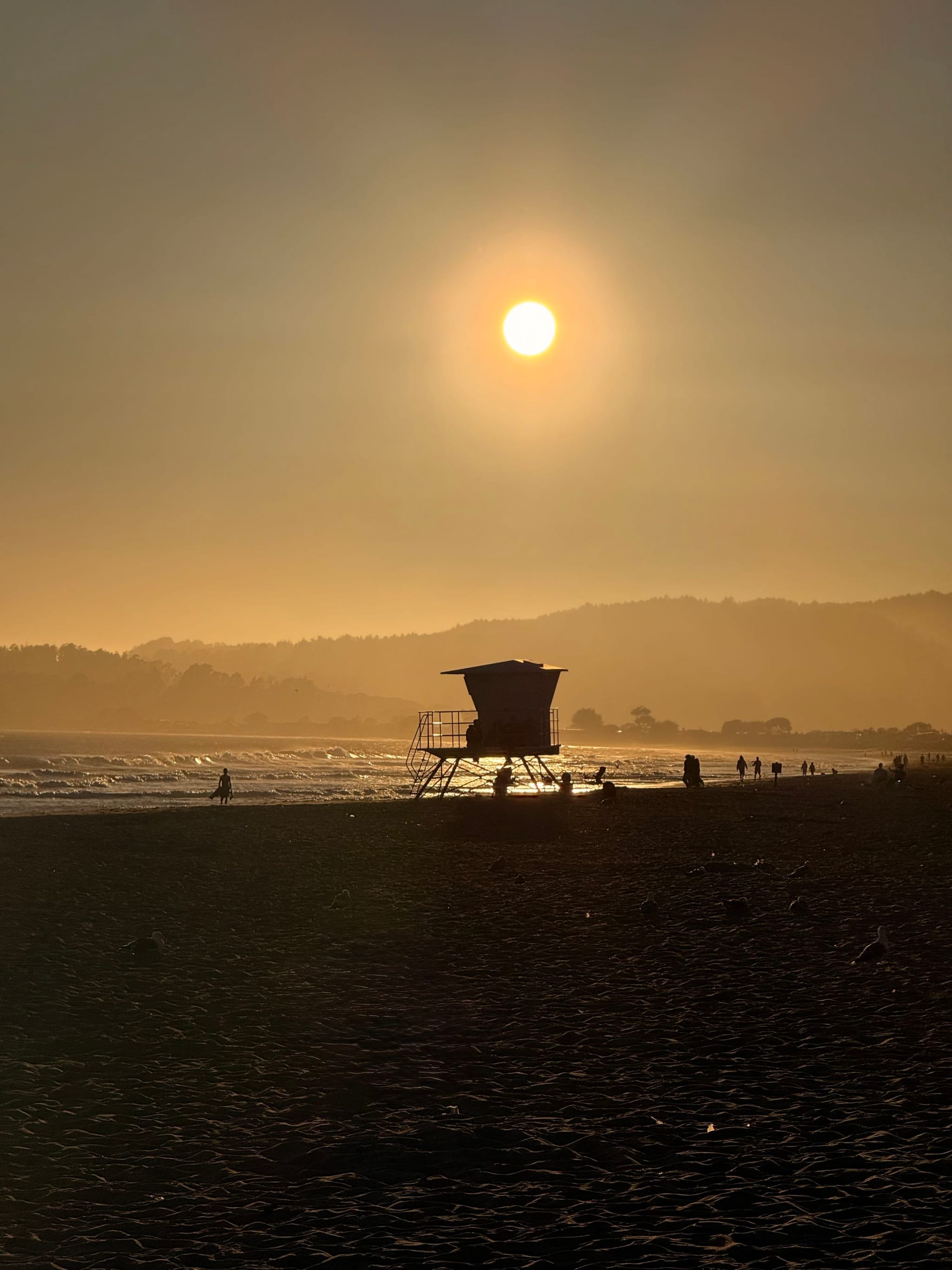 A lifeguard tower silhouetted against a sunset on a beach, with the sun low in the sky and hills in the background