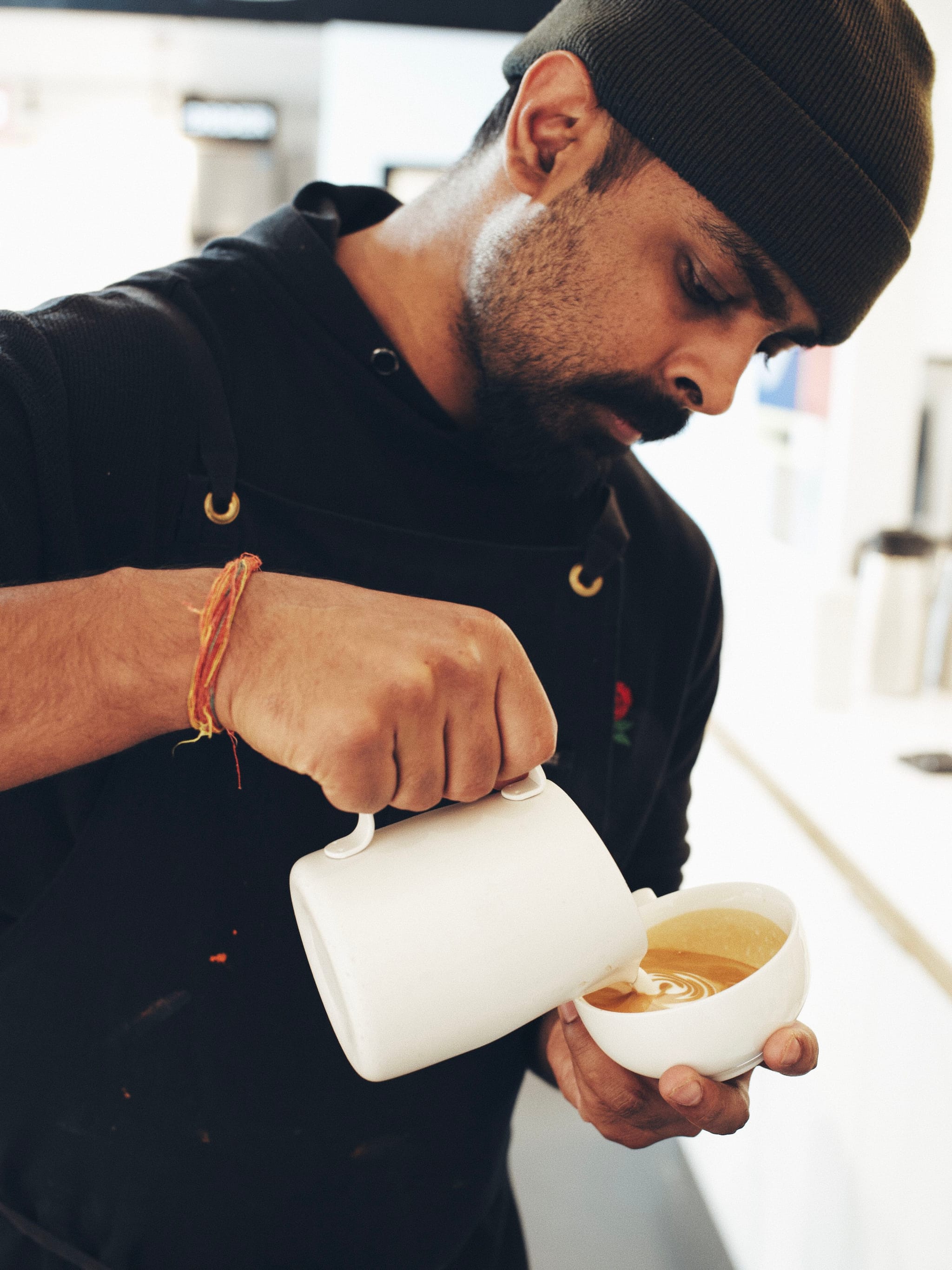 A person wearing a beanie and dark clothing is skillfully pouring milk from a jug into a cup, likely making latte art