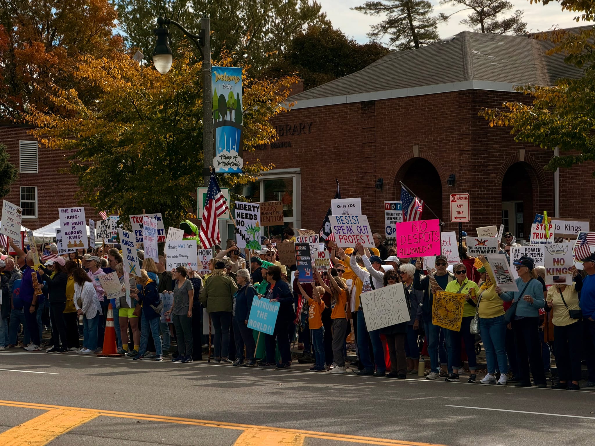 A large crowd lines a sidewalk in front of a brick building, holding protest signs and American flags on an autumn day