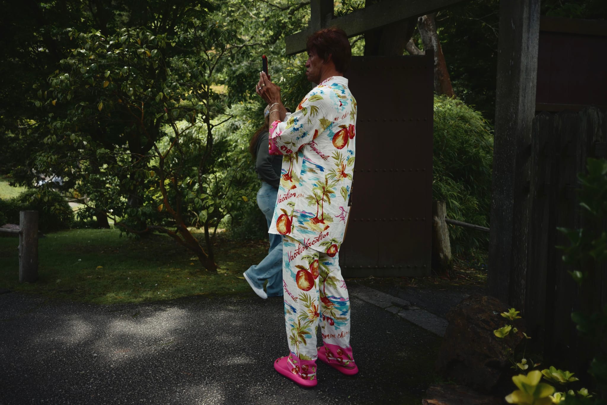 A person wearing colorful floral pajamas and pink shoes stands outdoors, holding a camera. The background features greenery and a wooden structure