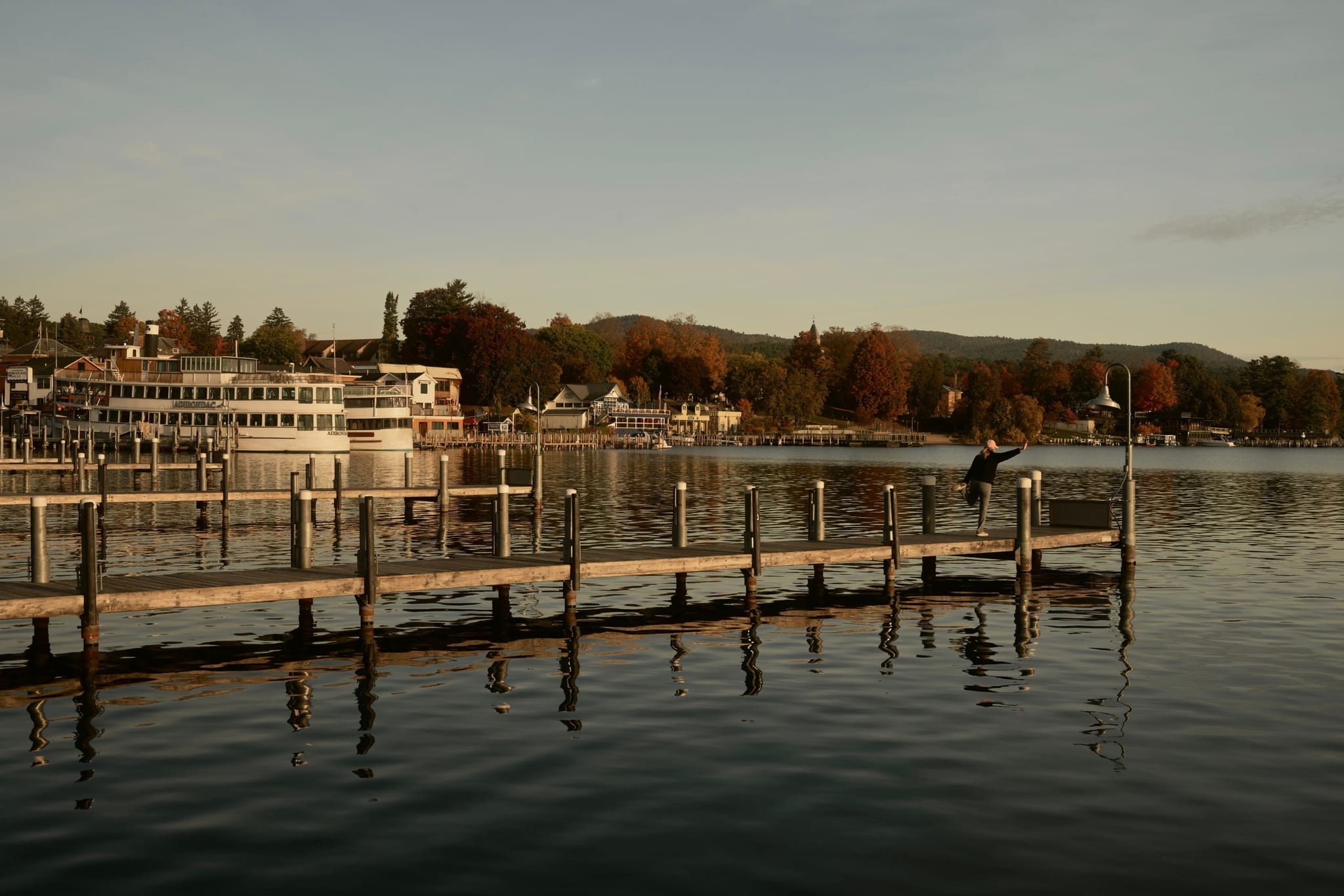 Wooden pier extending into a calm lake at sunset, with a moored ferry and lakeside buildings backed by autumn-colored hills