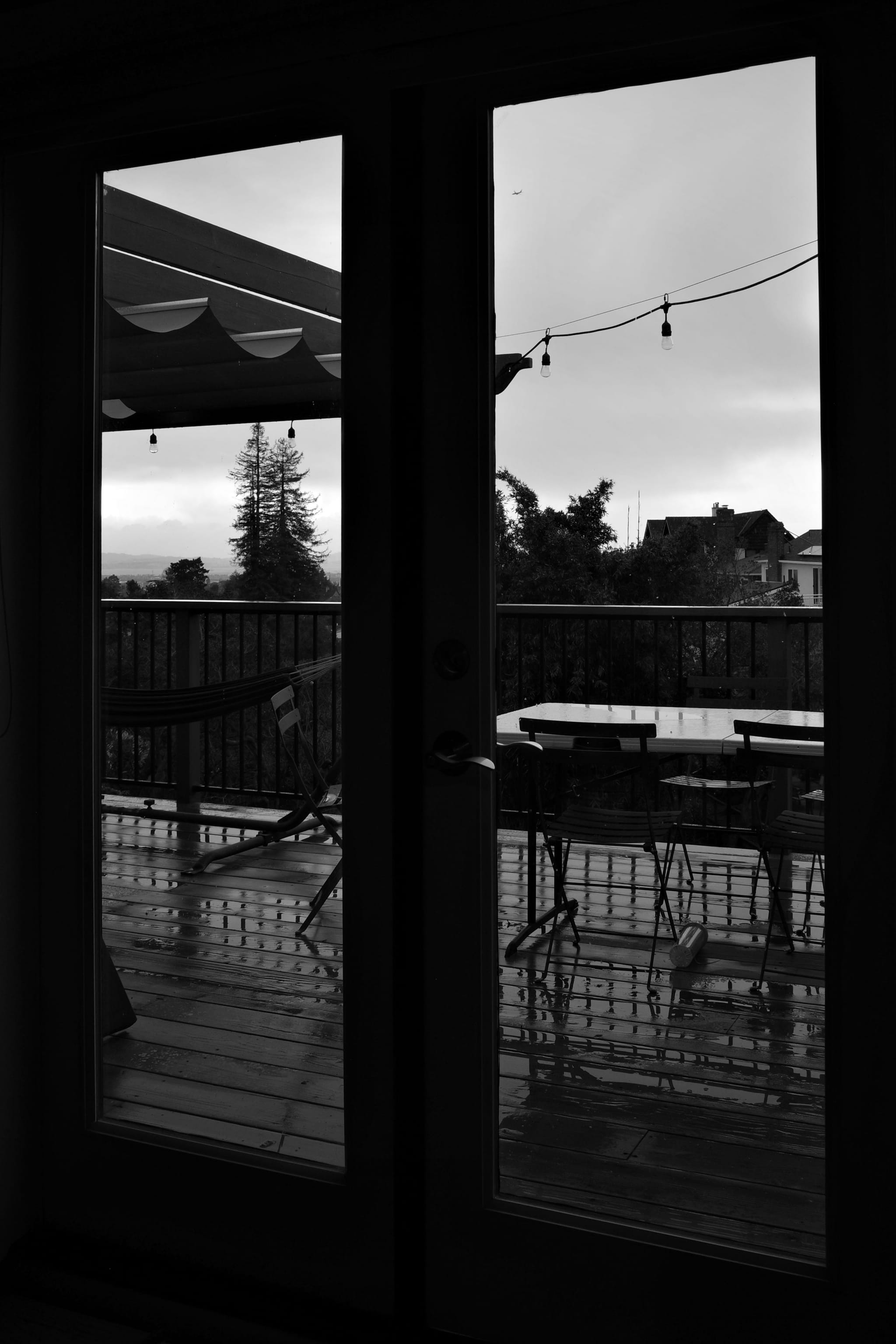 A black and white view through glass doors onto a patio with outdoor furniture, string lights, and a distant landscape