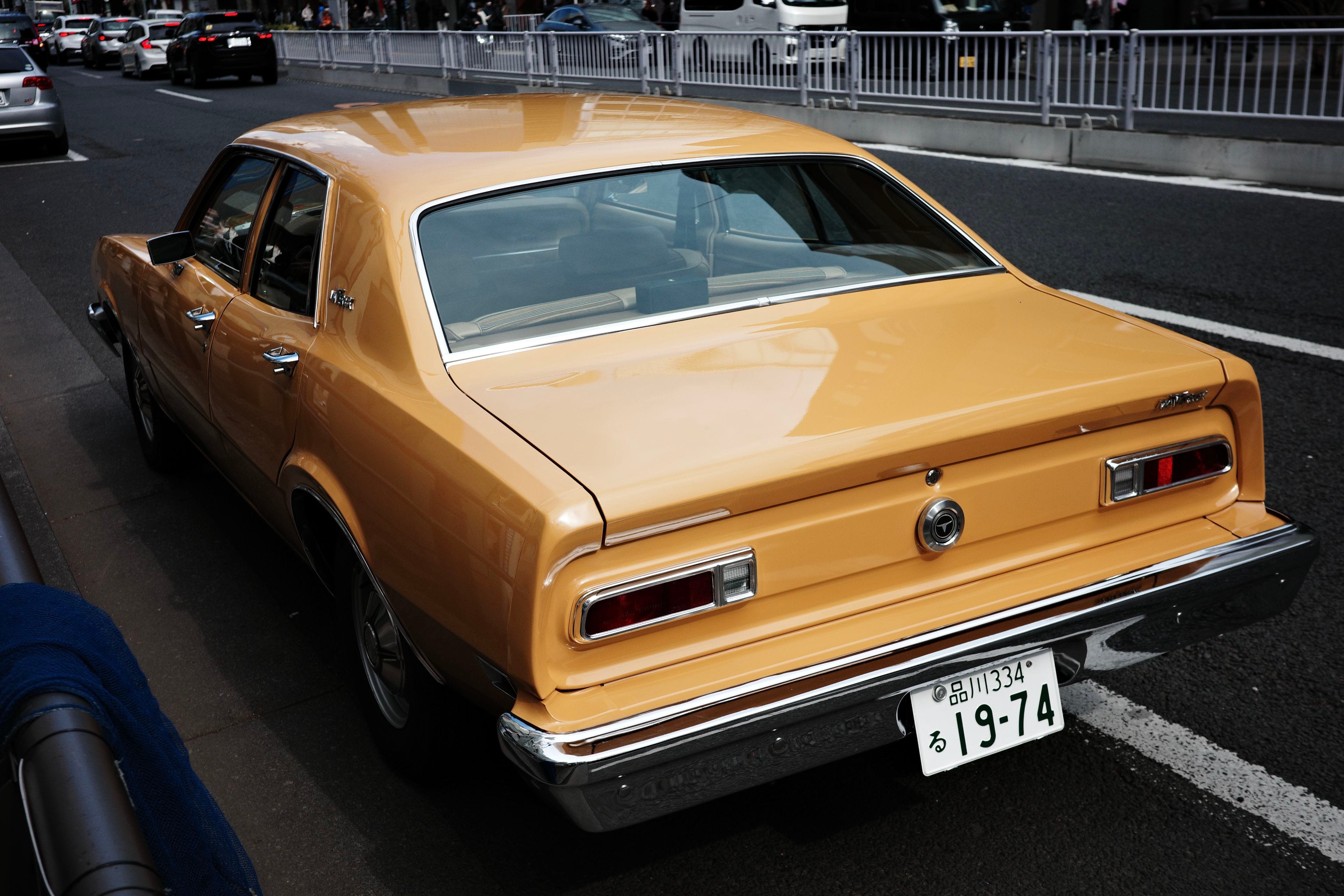 A yellow vintage car is parked on the side of a street, with a visible license plate and other vehicles in the background