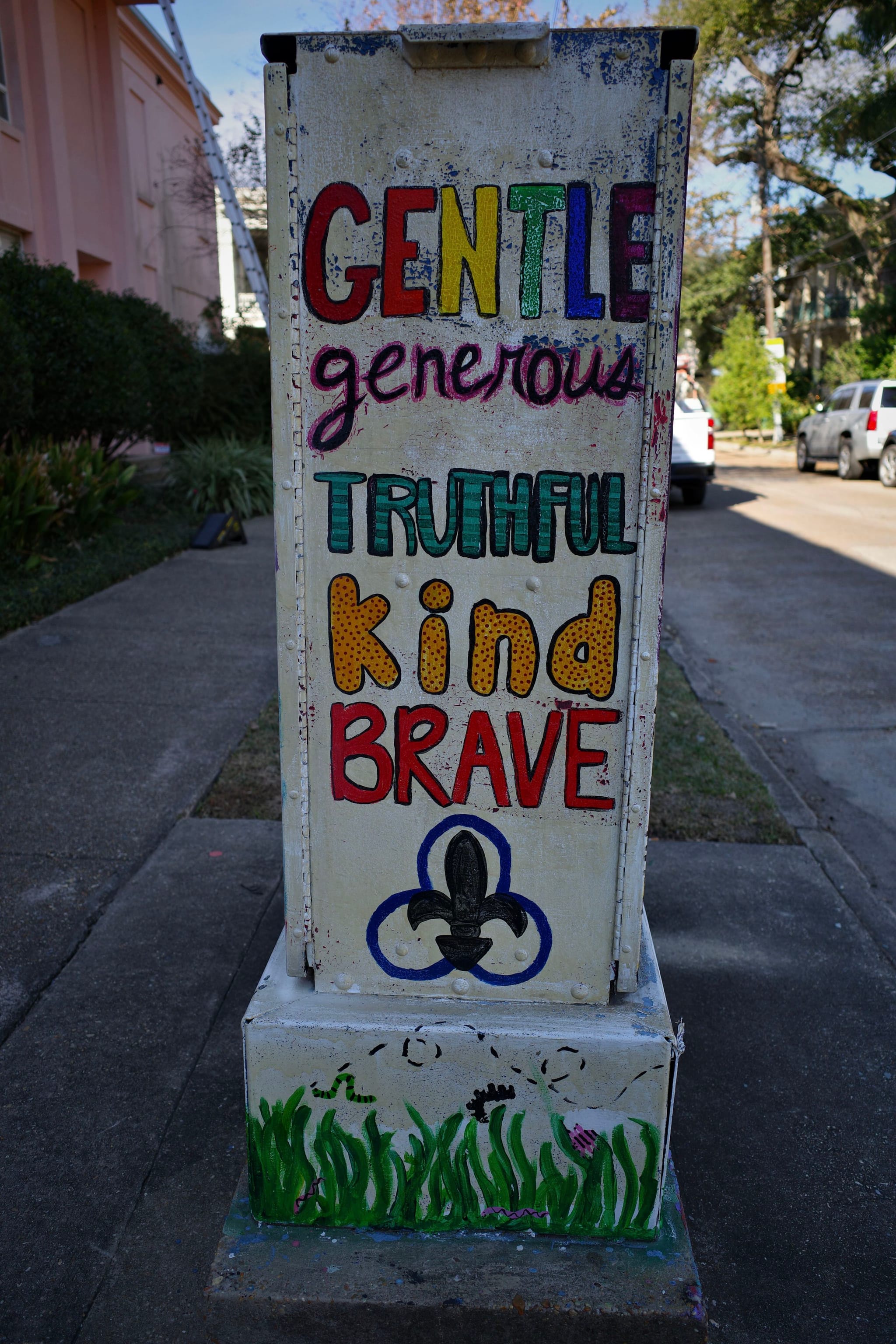 A painted utility box with the words GENTLE, generous, TRUTHFUL, kind, BRAVE in colorful letters, featuring a fleur-de-lis symbol and grass at the bottom