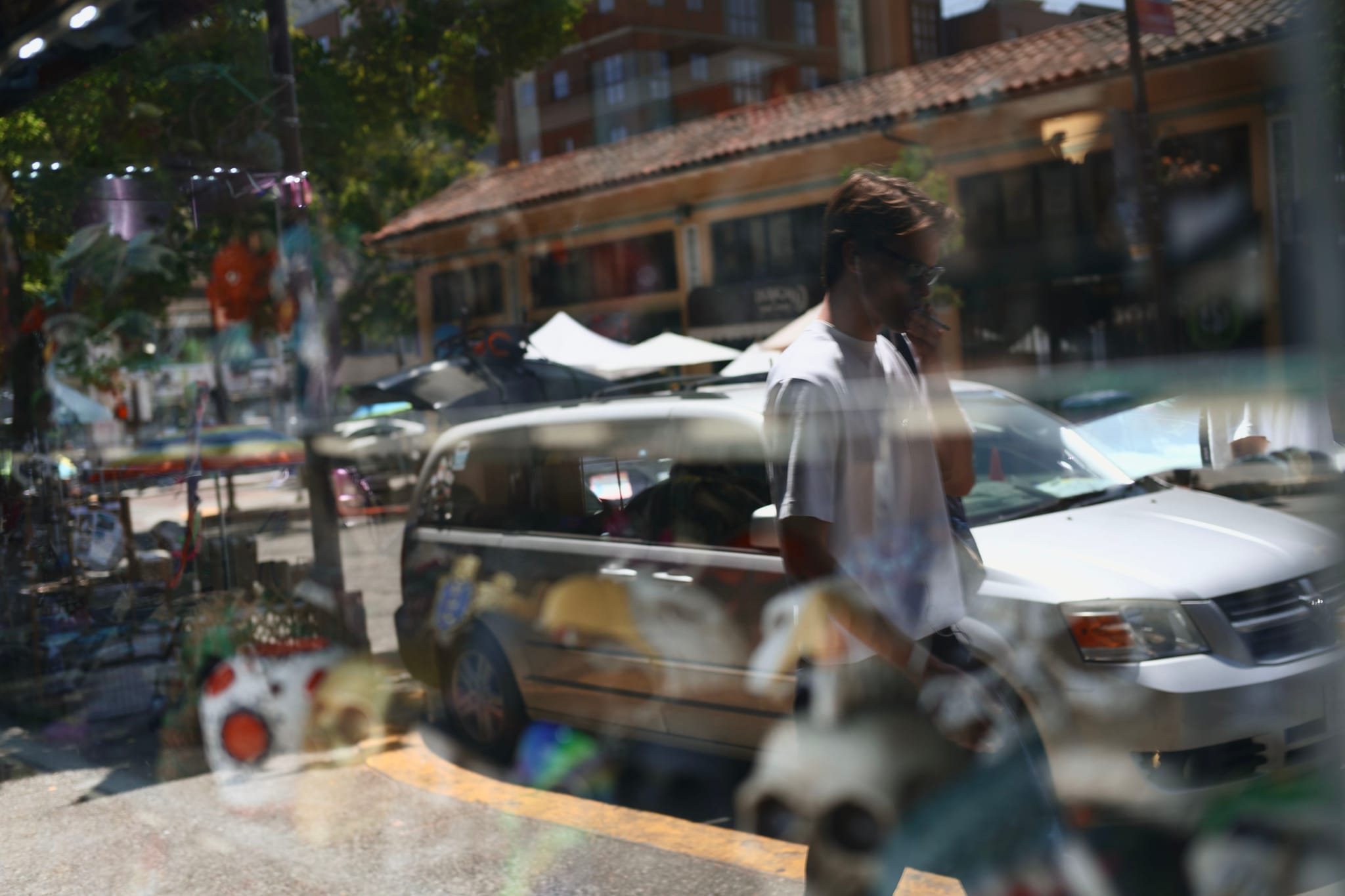 A person walking past a parked car on a sunny street, with reflections of various objects in a window