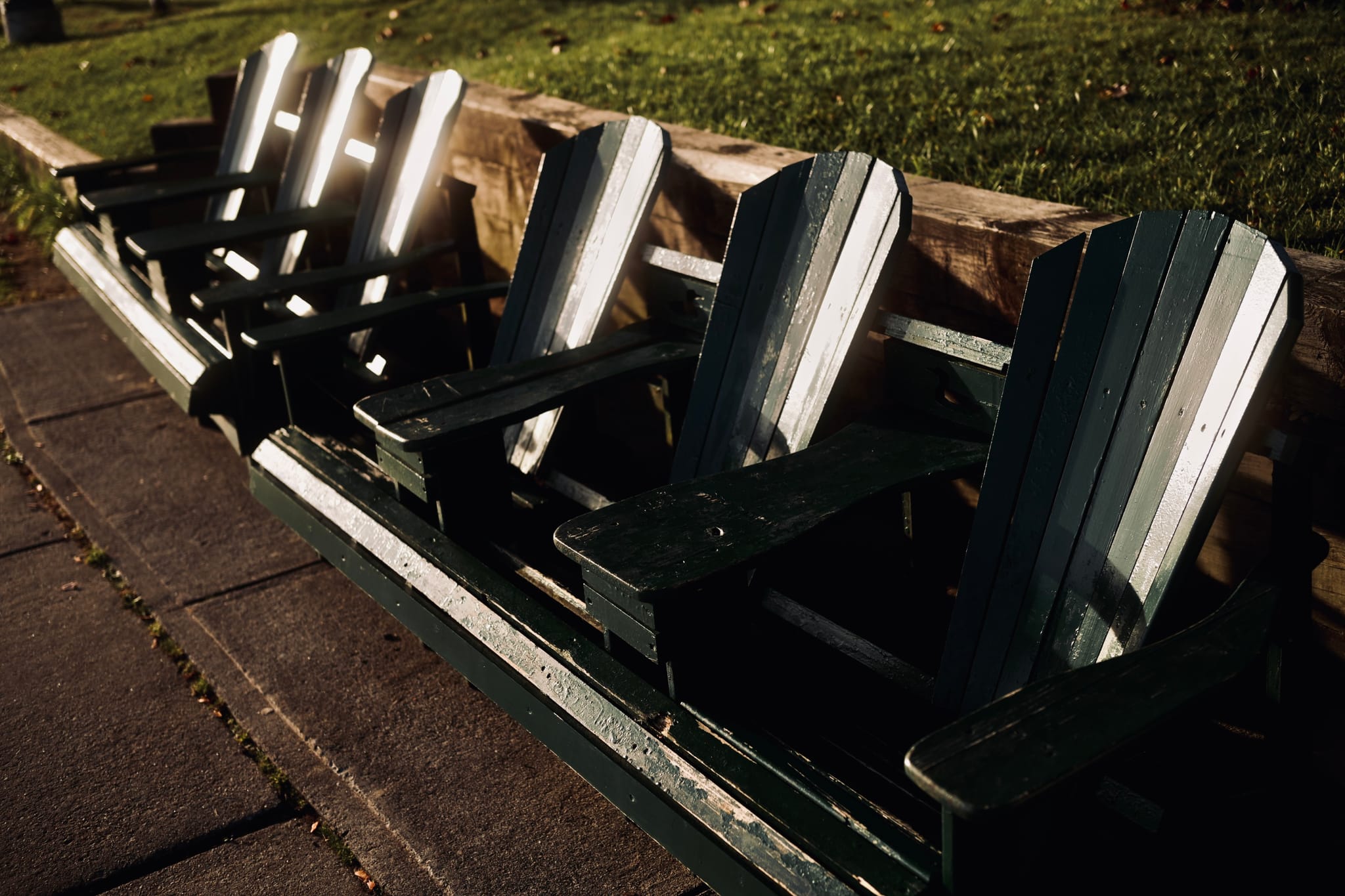 A row of dark green Adirondack chairs on a paved path beside grass, lit by low afternoon sun