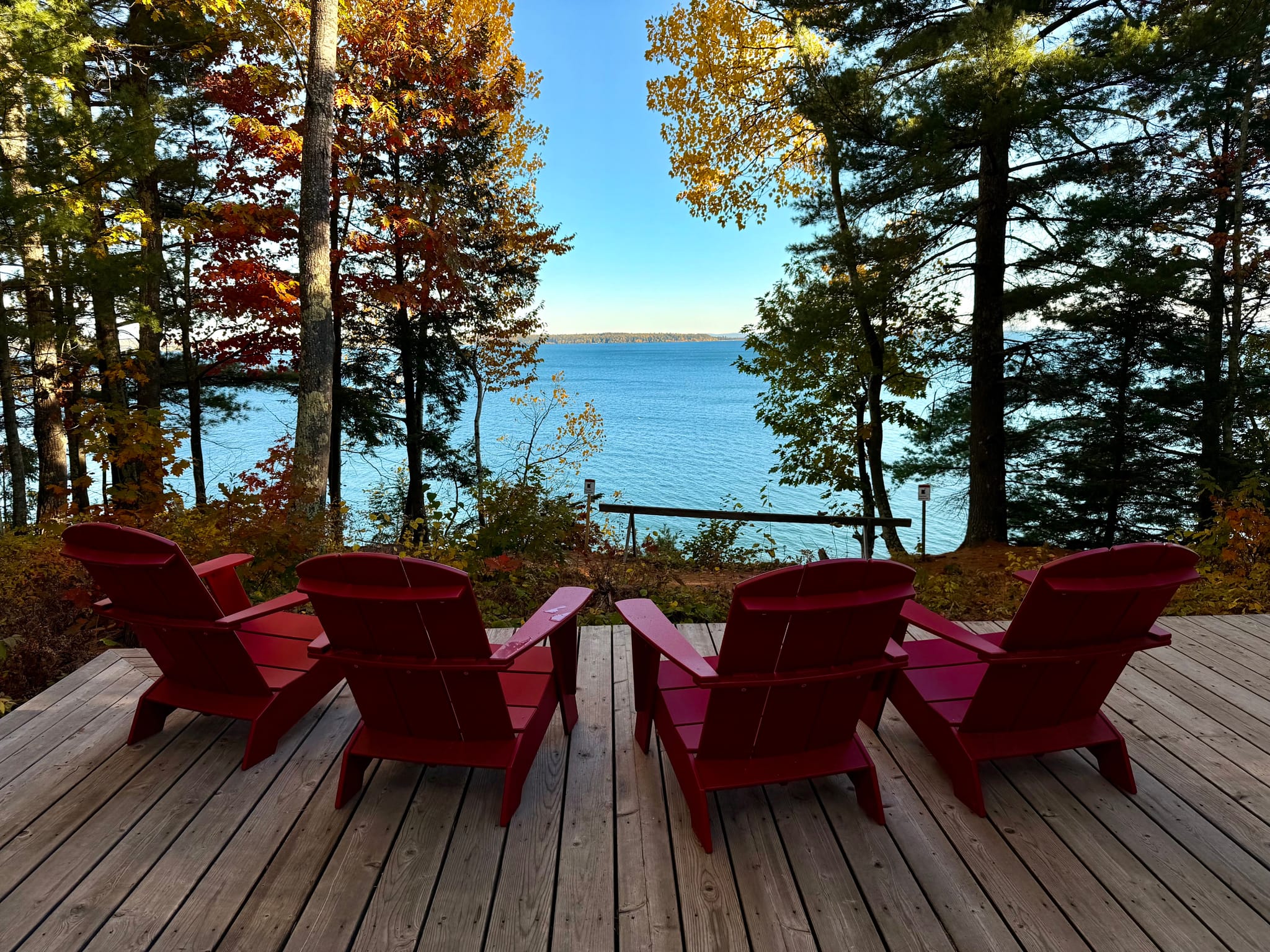 Four red Adirondack chairs on a wooden deck facing a calm lake, framed by tall trees with early autumn foliage