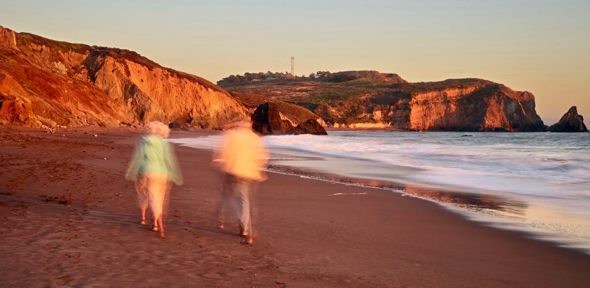 Two people walking along a beach at sunset with cliffs in the background