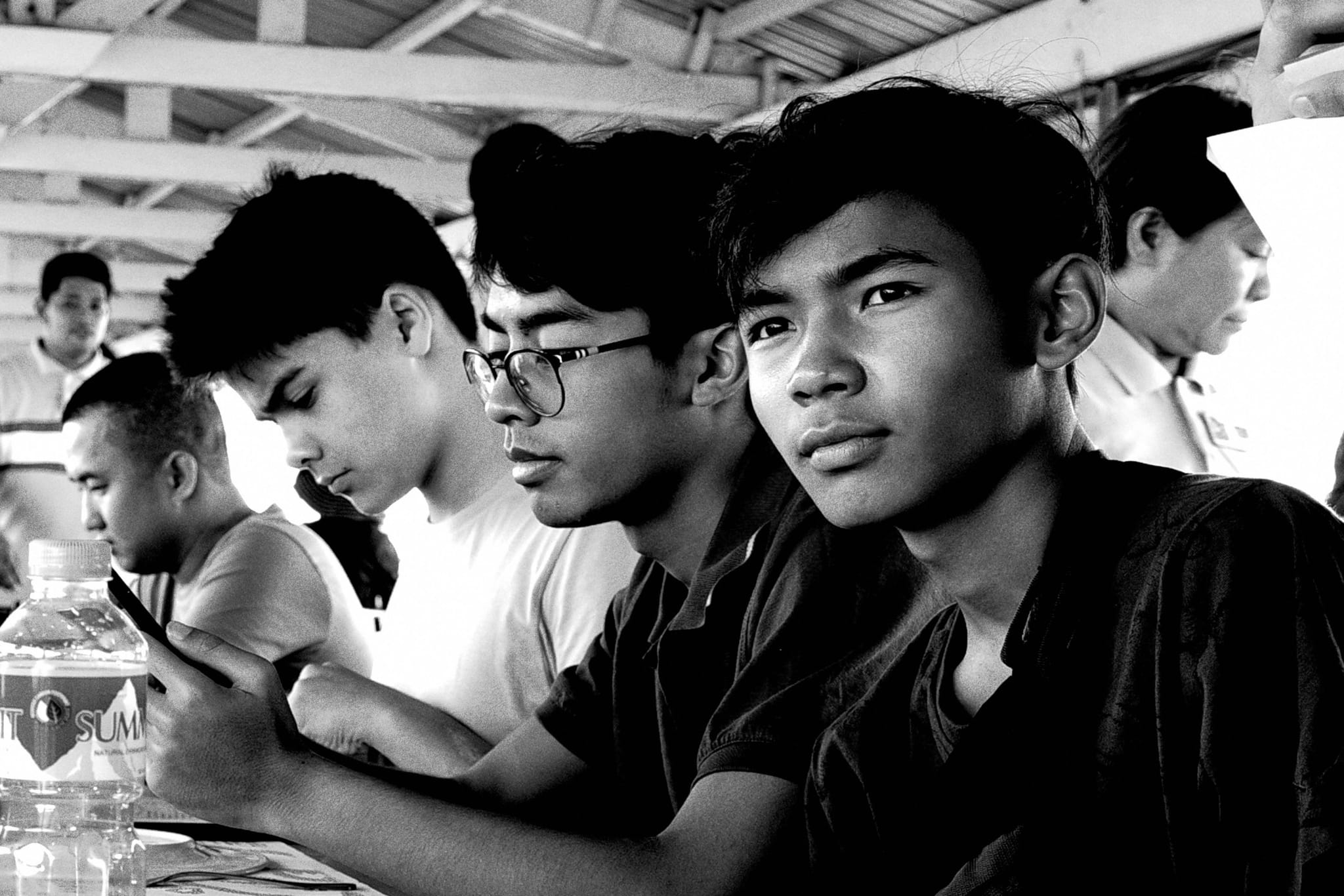 A group of young men sitting closely together, some looking at their phones, in a black and white setting