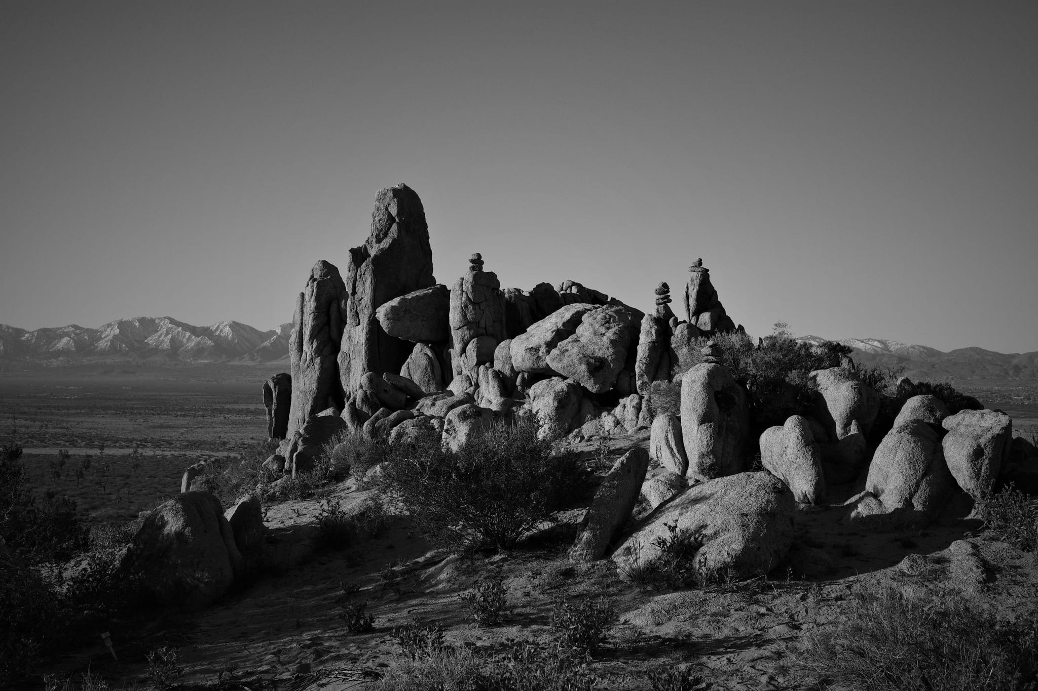 A black and white landscape featuring a rocky outcrop with variously shaped boulders in the foreground and distant mountains under a clear sky