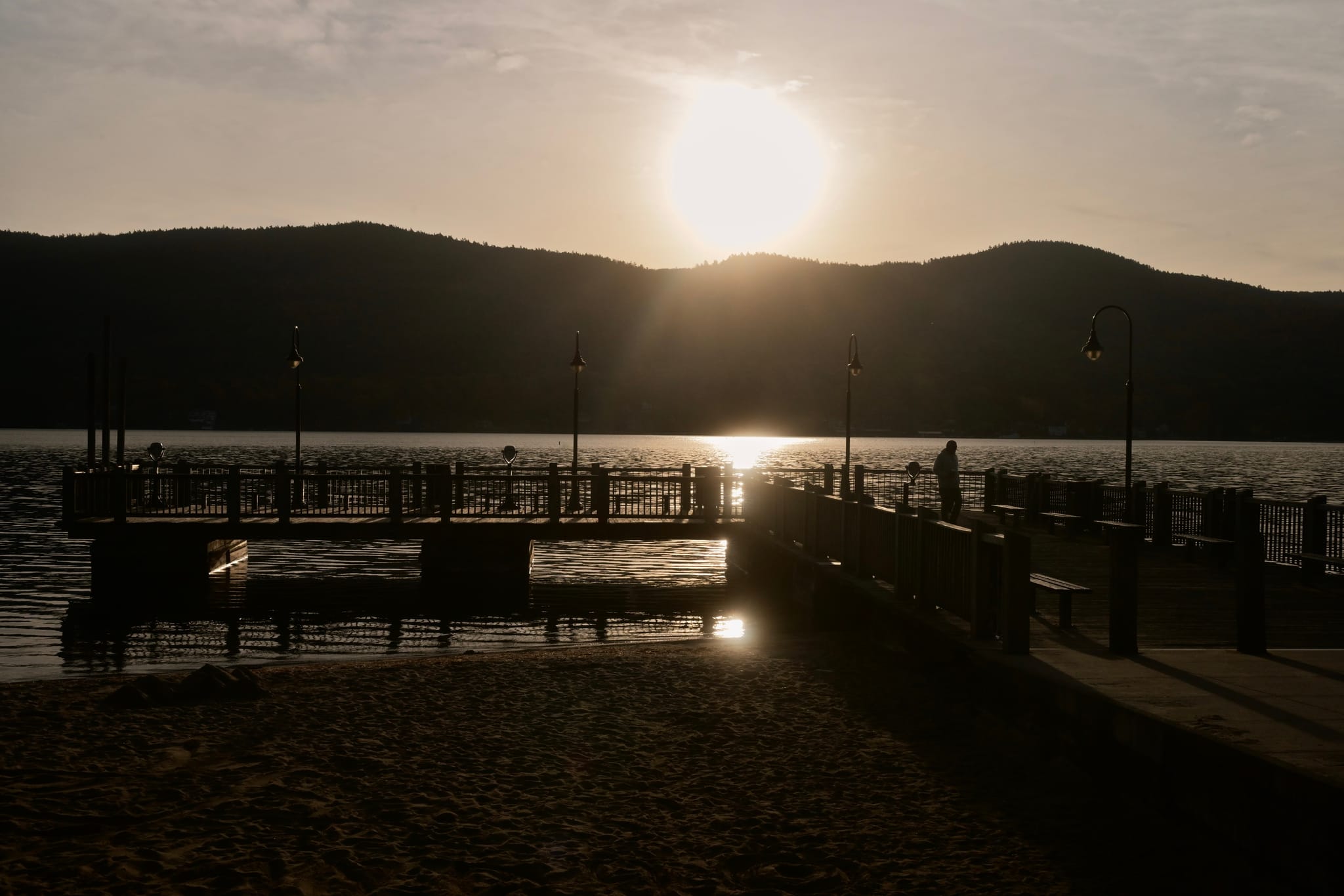 Sunset over a lakeside pier with silhouetted people and lampposts, mountains in the background, and sunlight reflecting on the water