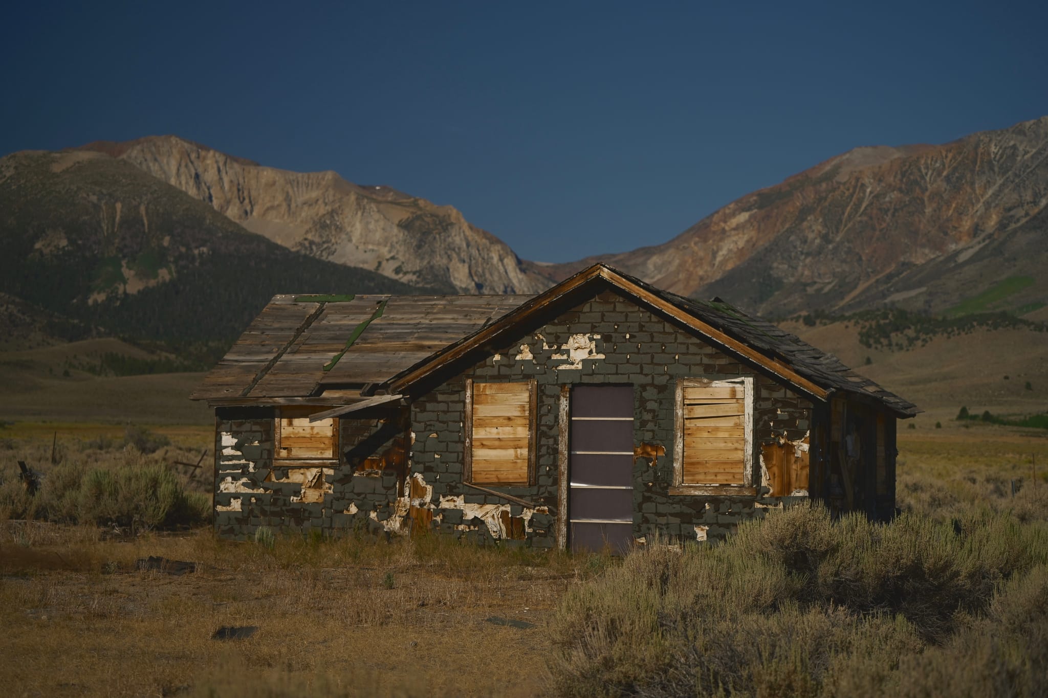 An abandoned, weathered cabin with boarded-up windows stands in a grassy field, surrounded by mountains under a clear blue sky