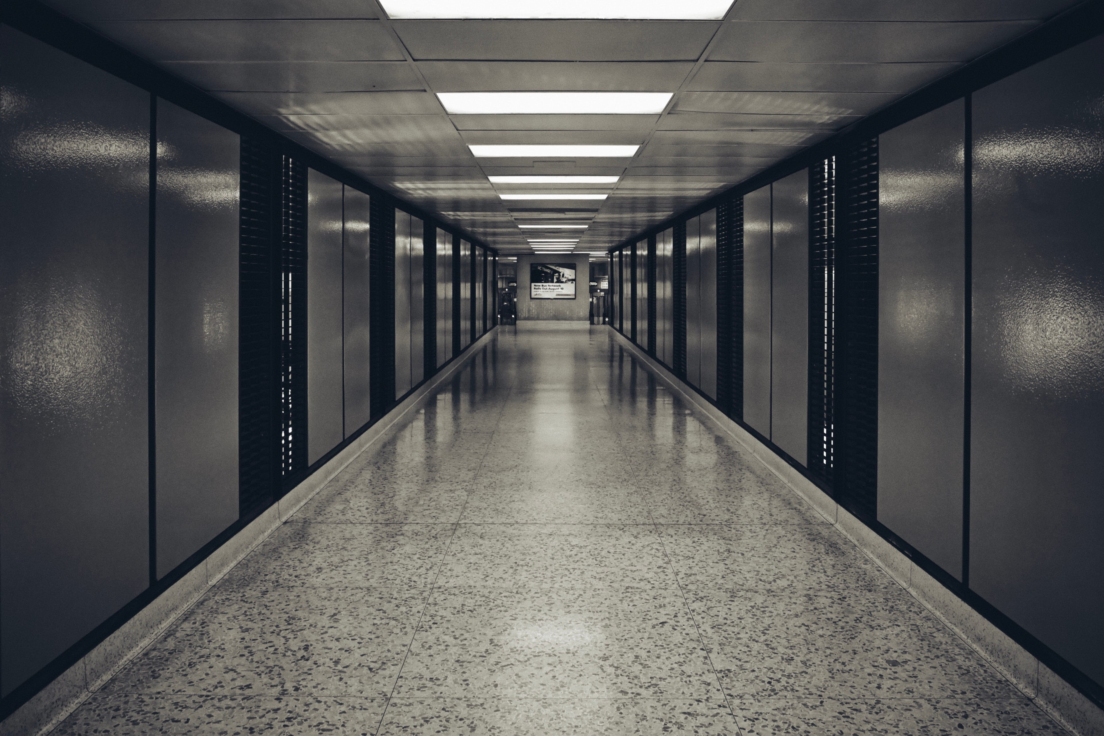 A long, empty corridor with a polished floor, flanked by dark walls and illuminated by overhead fluorescent lights
