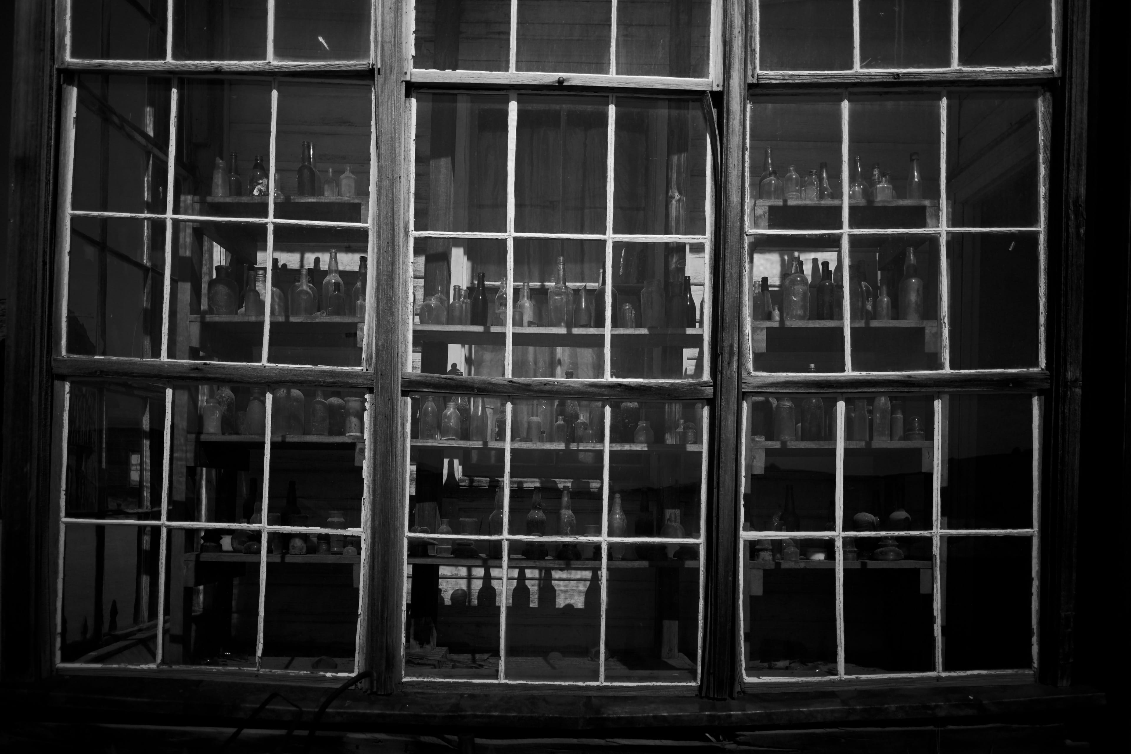 A black and white view through a window with multiple panes, revealing shelves filled with various bottles inside a dimly lit room