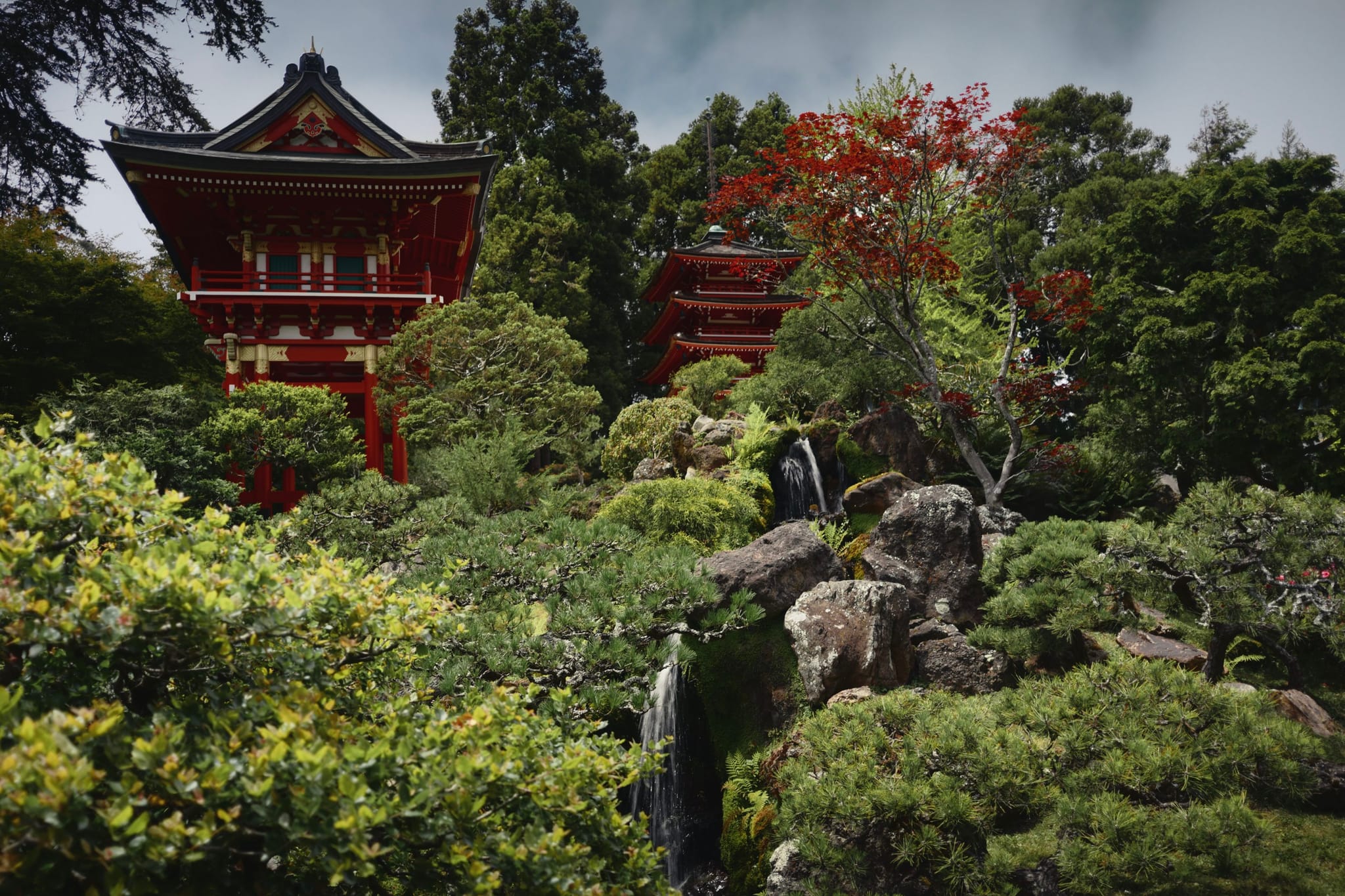 A traditional Japanese garden with lush greenery, a small waterfall, and a red pagoda-style building in the background