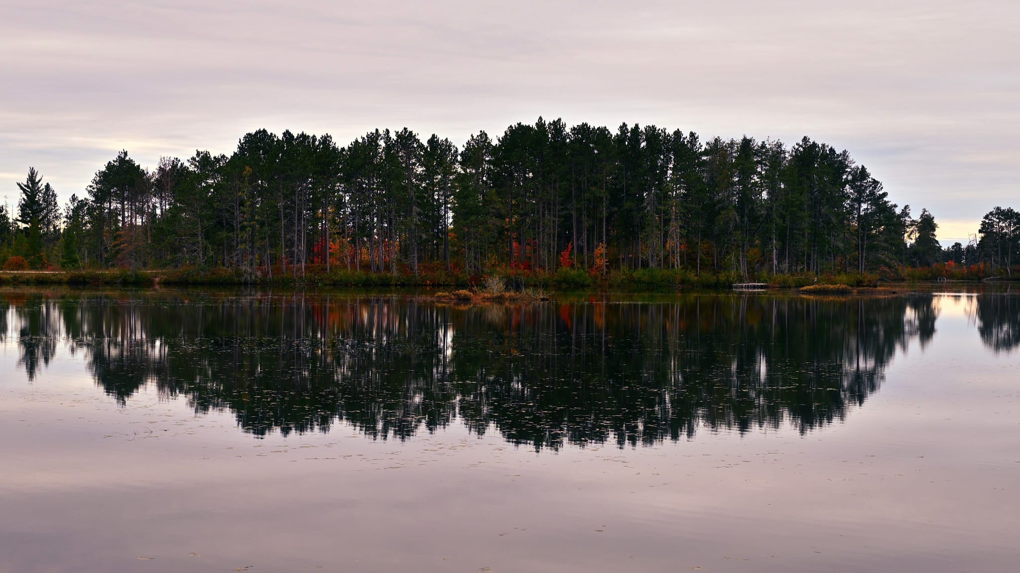 Still lake reflecting a tree-lined shoreline with autumn colors beneath an overcast sky