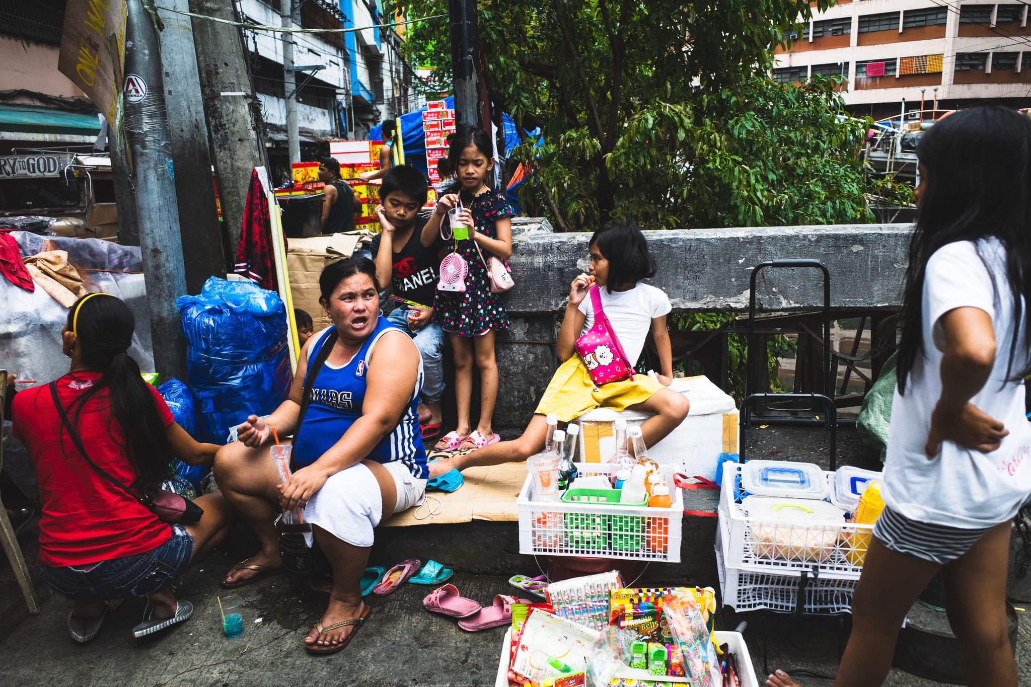 A group of people, including children, are gathered on a sidewalk with various items and boxes around them. Some are sitting, while others are standing, in an urban setting with buildings and trees in the background
