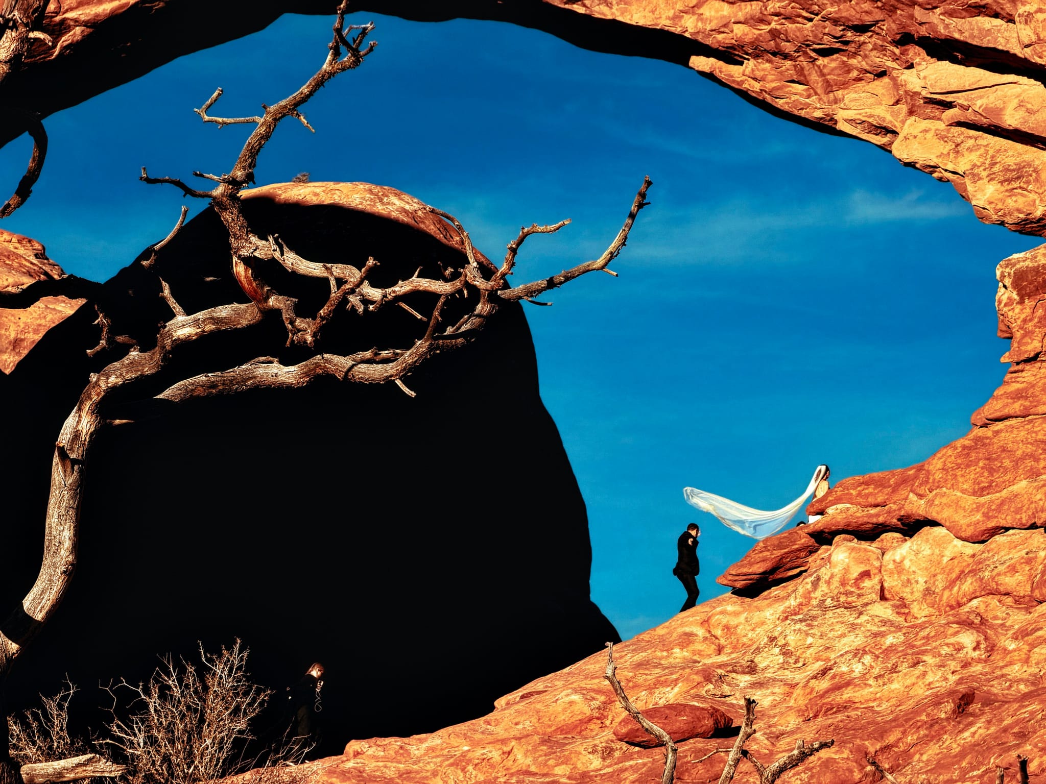 Bride and groom beneath a sandstone arch, veil flowing against deep blue sky