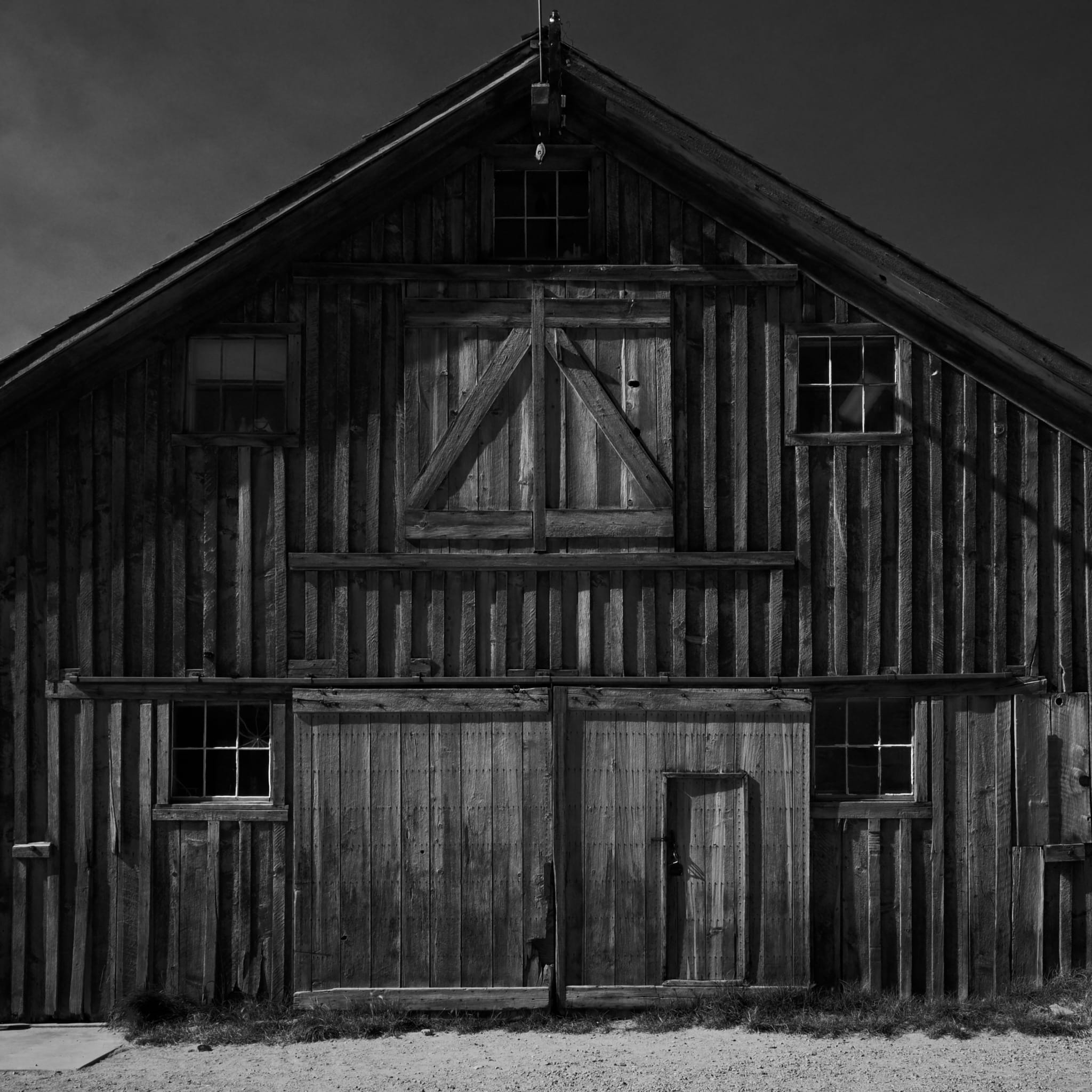 A black and white photograph of a rustic wooden barn with a gabled roof, featuring large double doors and several small windows