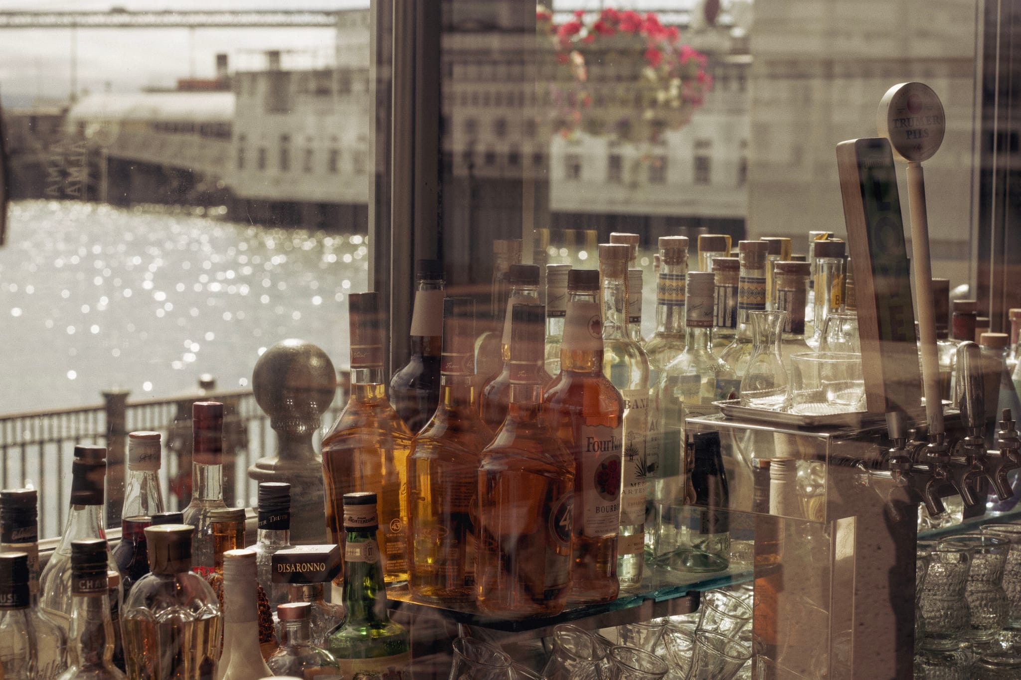 A collection of liquor bottles and bar taps is reflected in a window, with a waterfront view in the background