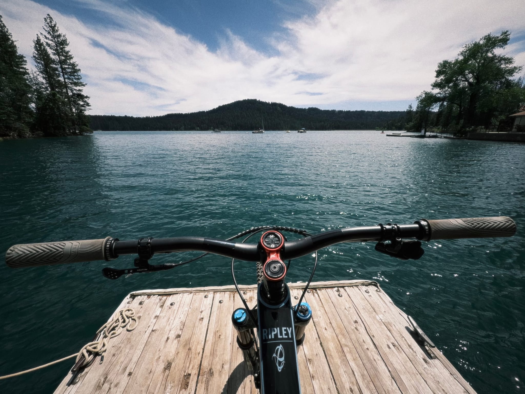 Handlebars of a mountain bike resting on a wooden dock facing a lake with forested hills under a cloudy sky